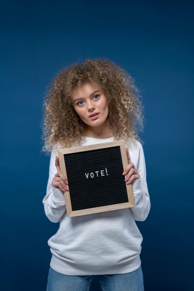 Curly-haired woman holding a letter board with 'Vote!' written, promoting civic duty.